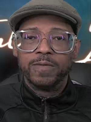 A medium shot of a man wearing a gray newsboy cap and clear-framed glasses. He has short, dark hair and a neatly trimmed goatee. The background is slightly blurred, with a hint of neon lighting. The man is looking directly at the camera with a neutral expression.