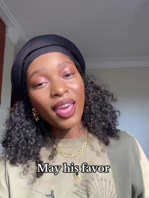 A woman with curly black hair wearing a black headwrap and a gold chain necklace is shown, against a soft, neutral background. The hook text "May his favor" is overlaid at the bottom. The text overlays on a casual outfit, looking directly at the camera with a slightly open mouth. The overhead lighting and simple background contribute to a clean and personal feel.