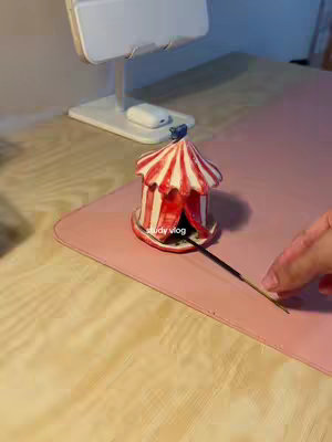 The image shows a desk setup. A light wooden surface forms the base, upon which sits a pink mousepad. A small, decorative red and white tent-shaped object is in the center, with an incense stick placed inside, trailing slight smoke. To the left, a white stand holds what appears to be a tablet or phone. A hand is reaching in from the right side, possibly tending to the incense stick.