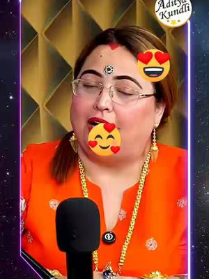 A woman with glasses, an orange top stands facing the camera. She has red lipstick and a bindi on her forehead. Her eyes are closed, and there are emoji stickers with hearts over her mouth and to the side of her head. In the background is a decorative gold pattern, and a microphone is in front of her. The image is framed with a purple and blue gradient border.