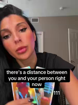 A woman with long black hair and dark makeup is holding tarot cards toward the camera. The image is framed from the shoulders up, with the woman's face in the center, looking directly at the viewer. A black bar with white text overlays the bottom of the image.