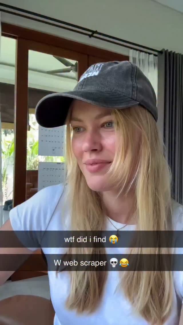 A woman with long blonde hair wearing a grey cap, and a white t-shirt. The background shows parts of a house.