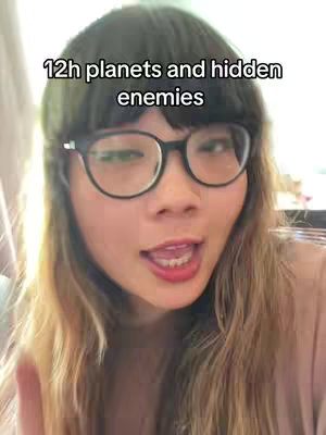 A woman with dark bangs and glasses is speaking directly to the camera. Text that reads '12h planets and hidden enemies' is overlaid on top of the image. 