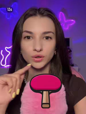 A woman with long brown hair is centered in the frame, looking directly at the viewer with a soft expression and open lips. She is lightly touching her index finger to her mouth. Behind her, a purple background features neon butterfly and other shapes, partially visible. She is holding a pink ping-pong paddle over a fluffy pink surface. The image has a soft, warm lighting.