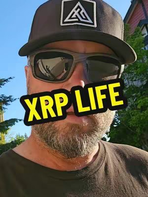 A selfie of a man wearing a black cap and sunglasses, with the text "XRP LIFE" overlaid in a yellow rectangular box. Behind him is a blurry background of greenery and a building with a red roof.