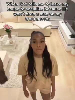 A selfie-style shot featuring a young Black woman standing in a well-lit living room. The central focus is on her face, with a slightly elevated camera angle. She is wearing a matching beige t-shirt and shorts set and her long dark hair is down. The background includes a light-colored sectional sofa, a glass coffee table with a vase of flowers, and a white rug.