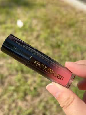 A hand holding a tube of lipstick, angled diagonally against a blurred green-and-brown background of foliage. The lipstick tube is cylindrical with a black cap and a clear bottom showing a pinkish-red color. The brand "PECOLOVERS" is printed in gold letters on the tube. The hand is holding the lipstick in a way where the fingers wrap around it firmly.