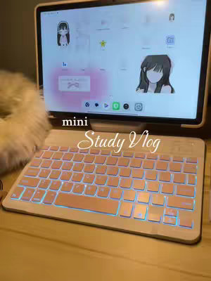 A shot of a lit-up keyboard and tablet displaying a screen with anime-style images. The keyboard is centered at the bottom, glowing with pink and blue light. Above it, a tablet showcases what appears to be a study app. The image suggests a study vlog setting.