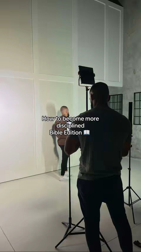 A behind-the-scenes shot of a video with two men, one of whom is the main subject. The main subject is wearing a brown shirt and black pants, standing in front of a white wall. The other man is visible from the back, wearing a dark shirt. A stand with a spotlight and camera equipment are set up, suggesting a filming location.