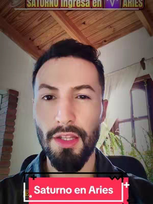 A man with a beard looks at the camera, with a backdrop that includes a wooden ceiling and a partial view of a window. A text overlay displays the words, "Saturno en Aries".