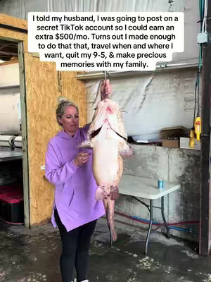 A woman stands in a slightly grungy, indoor space. She is holding up a large, light-colored fish, suspended in front of her. She is wearing a purple hoodie and black leggings, and the background includes a wooden wall and a metal door frame. A table and a can are on the side, adding to the casual setting.