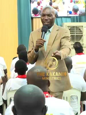 A man in a tan suit stands in front of an audience, holding a microphone and a sign with the text "KM THE KAKANDE MINISTRIES". The image is well lit and shows the man speaking. The background is slightly blurred, with more audience members visible. The color palette is natural and consistent with an indoor setting.