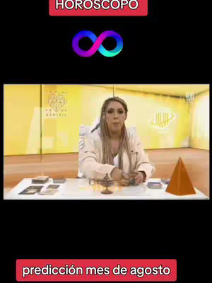 The cover image for the video has a black background. In the top center is a colorful infinity symbol. Below it, a woman is seated, with blonde hair, looking towards the camera. She is in a studio setting and is seated behind a desk with various items on it; cards, a pyramid, and a golden item. There is text at the top "HOROSCOPO" in red and a banner at the bottom "predicción mes de agosto" also in red.