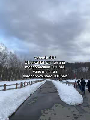 The image shows a winter scene with a road or path leading into the distance, flanked by banks of snow. A text overlay provides a Bible verse in Indonesian. The sky is overcast.