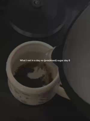 The image shows a monochrome overhead shot of a cup being filled from a pitcher. The cup is white, and the liquid is dark, possibly coffee. Text overlays the cup, describing the content as "What I eat in a day no (processed) sugar day 6".