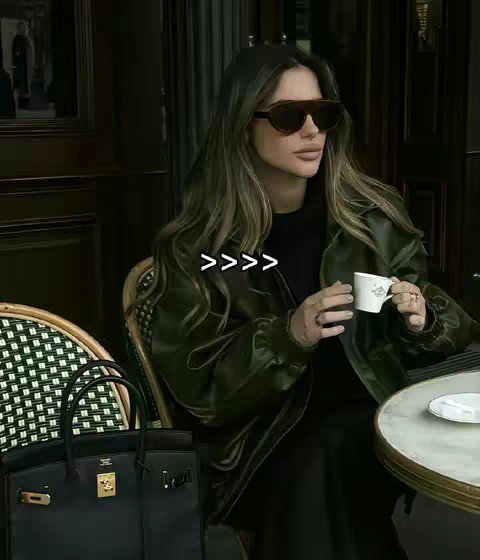 A woman in sunglasses and a green jacket sits at an outdoor cafe table, holding a cup. A black handbag rests on the ground beside her. The background is blurred, suggesting a cafe setting.
