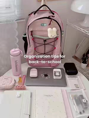 A top-down shot featuring a neatly organized desk and items related to studying. A pink backpack is central, with a notebook, a laptop, water bottle and stationary items placed on the table. The color palette is soft and girly, with pinks and whites dominating, exuding a sense of calm and preparedness.