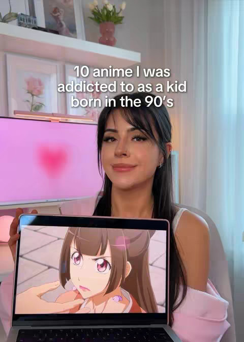 A woman is holding a laptop with an anime scene on the screen. The background includes a white shelf with decorations, a pink-tinted computer monitor, and a window with a curtain.