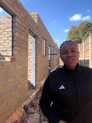 A woman in a black Adidas jacket is standing in front of a brick building under construction. The background is a blue sky with some clouds and a fence.