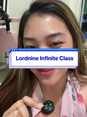 A selfie-style shot featuring a young woman with light-brown hair. She is in close proximity to the camera, and a digital-style text overlay labeled "Lordnine Infinite Class" partially obscures her face. She appears to be holding a small, black device with green lights, suggesting she is interacting with it. The background is slightly blurred, with a hint of a room setting including some furniture.