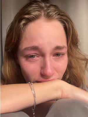 A close-up shot of a person's face, likely a young woman, with visible signs of crying. Her eyes are red and tear-filled, and she is looking down, resting her chin on her left forearm, with her hand covering part of her face. A silver bracelet adorns her wrist. The lighting appears to be soft and diffused, with a neutral background of soft grey. The overall tone is one of sadness and vulnerability.