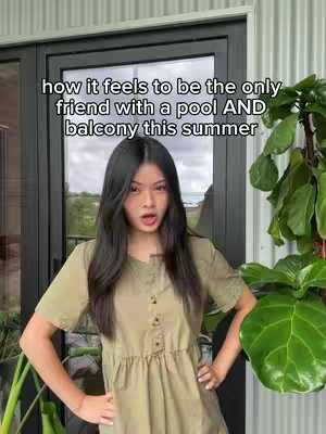 A woman with long black hair stands in front of a glass door with a verdant background. She wears a button-down dress and has her hands on her hips with an open mouth expression. The overlay reads 'how it feels to be the only friend with a pool AND balcony this summer'.