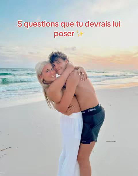 A couple embraces on a beach with a sunset as the backdrop. The woman is in white pants, and the man is shirtless with black shorts. Text overlayed reads '5 questions que tu devrais lui poser'.