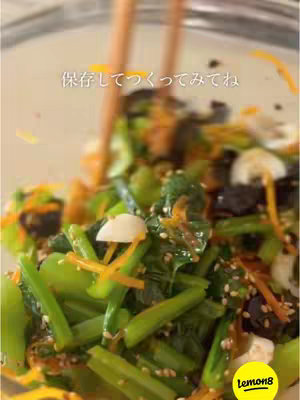 Close-up shot of prepared food being mixed inside a clear glass bowl. The dish contains green leafy vegetables, orange julienned carrots, black pieces of a dried seaweed and white ingredients, possibly from the chives. Two wooden chopsticks are mixing the ingredients. The image is brightly lit, with the background out of focus.