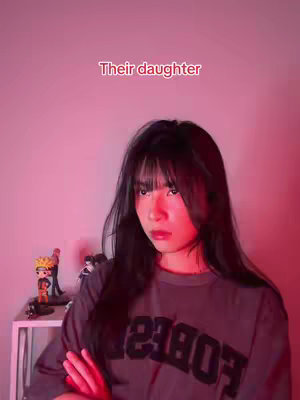 A young woman with black hair and bangs is the focus of the image. She wears a gray t-shirt with bold black text. The background is a solid pink color, with a small statue of a character on a white shelf on the left side. The words "Their daughter" appear in red text at the top.