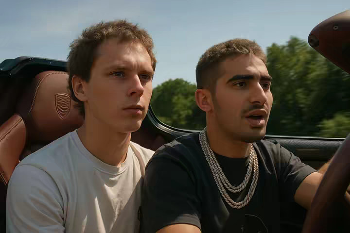 The cover image showcases two young men inside a car, likely a convertible, with the top down. The man on the left is wearing a white t-shirt and appears to be looking off to the side with a neutral expression. The man on the right has a more animated expression, with his mouth slightly open as though he is mid-sentence, and is wearing a black t-shirt and gold chain. They are set against a background of a sunny sky and green trees, suggesting a drive on a pleasant day. The car's interior is visible, providing context to the setting.