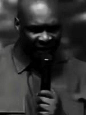 A black and white, slightly blurred image of a man holding a microphone. He is wearing a collared shirt and appears to be speaking. The background is out of focus.