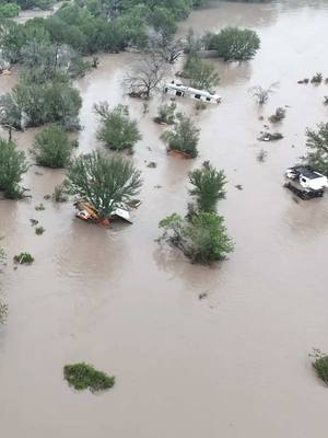 An aerial view shows a flooded area with brown, churning water. Trees and bushes dot the landscape, partially submerged. Several structures, possibly RVs or small buildings, are barely visible, some half-submerged. The overall composition is chaotic, conveying the devastation of a flood.