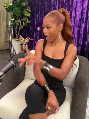 A woman with red hair in a high ponytail sits in a chair, gesturing with her hands and mouth open as if speaking with a surprised expression. She wears a black dress and large metallic cuffs. In the background are purple drapes and a microphone, suggesting a podcast or interview setting.