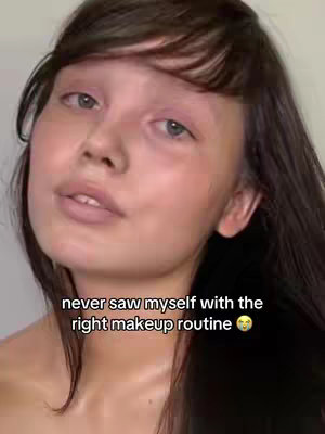 A selfie-style shot with a young woman looking at the camera. The background is blurred and neutral. The woman has brown hair with bangs, light skin, and is wearing some sort of makeup, the text overlay reads 'never saw myself with the right makeup routine'.