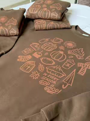 A brown sweatshirt laid out with a fall-themed design. The design includes various sketched images such as pumpkins, leaves, and a pie with the text "IN EVERY SEASON, GOD IS GOOD!" in the center. Above are folded brown sweatshirts or blankets.