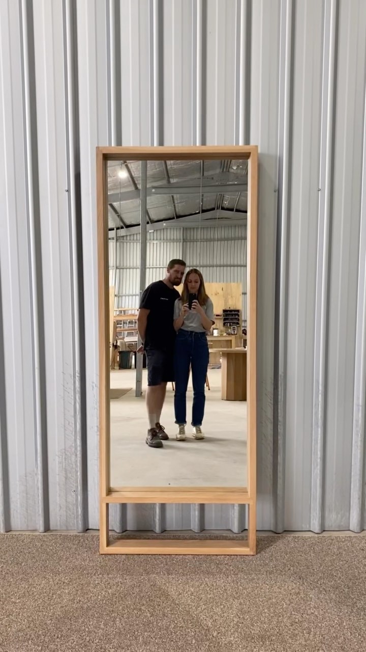 A full-length mirror reveals a man and woman standing inside a workshop. The man is on the left, wearing a black t-shirt and shorts, while the woman is on the right, wearing a grey t-shirt and jeans. They appear to be taking a selfie, with the woman holding a phone. The background includes a metallic wall and equipment, suggesting a workspace. The mirror is framed with wood.