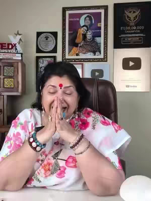 A woman with her hands clasped in prayer, eyes closed, is the central focus. She is wearing a floral top and adorned with jewelry, including bracelets and a bindi. Behind her is a wall decorated with framed pictures, awards, and two YouTube play buttons