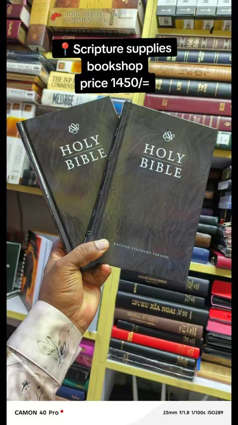 A close-up shot features a hand holding two black Holy Bibles, with the spines facing towards the viewer. The text 'HOLY BIBLE' is prominently displayed in white against the black cover. The background reveals bookshelves filled with a variety of books, including some with colorful spines. At the top left, a text overlay labels "Scripture supplies bookshop price 1450/=", with a red location pin icon next to the text. The scene is lit with natural light, creating shadows and highlights on the books and the hand.