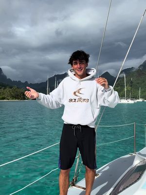 A young man stands on a boat, smiling with his arms open. He wears a white hoodie, black shorts, and casual shoes. The background includes a turquoise sea, a cloudy sky, and distant islands and sailboats.