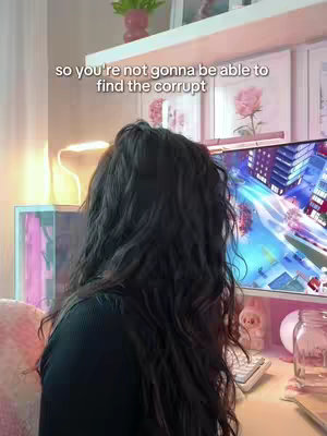 A person with long dark hair is the focus, seen from behind, seated at a desk. A computer monitor displays a colorful city, and text overlay appears atop the image. The desk has a keyboard, and the room contains a lamp and decorative items.