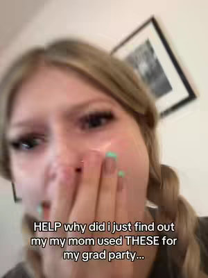 A close-up shot of a person, likely female, with blonde hair pulled back and green painted fingernails looks directly at the camera with a distressed expression, her hands covering her mouth. The background is blurred, suggesting an indoor setting with a picture frame visible.