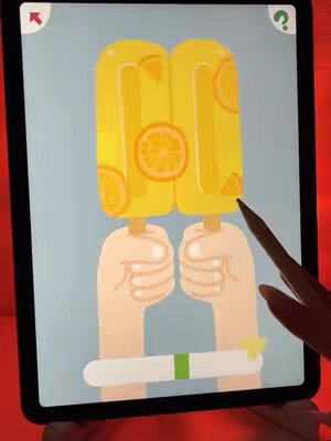 The cover image shows a tablet screen featuring a mobile game. The game displays two stylized, yellow popsicle illustrations, each adorned with orange slices, held by a pair of hands rendered in a simple, cartoonish style. The background is a light blue. A hand with a stylus suggests user interaction, completing the scene. The overall look is clean and inviting.