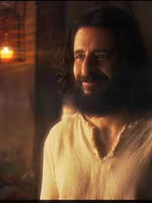 The image is a medium shot featuring a man with long, dark hair and a full beard, dressed in a simple, off-white tunic. His face is turned slightly, exhibiting a gentle smile and eyes that gaze to the side. The background, blurred but visible, suggests a warmly lit interior setting, indicating a location with soft light, perhaps a candle or a lamp, which complements the man's complexion.