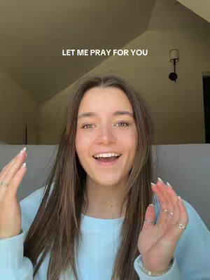 A young woman with long brown hair looks directly at the camera with an open smile. She's wearing a light blue long-sleeved shirt. Her hands are raised, palms facing forward, suggesting an inviting or welcoming gesture. The text overlay reads, "LET ME PRAY FOR YOU". The background is a soft, neutral beige, possibly a ceiling or attic space with a light fixture visible. Overall, the image has a warm, inviting feel, suggesting a message of hope and support.