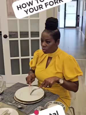A woman in a yellow dress sits at a table set for dining, demonstrating how to hold and pivot a fork. A text overlay reads, "HOW TO PIVOT YOUR FORK."