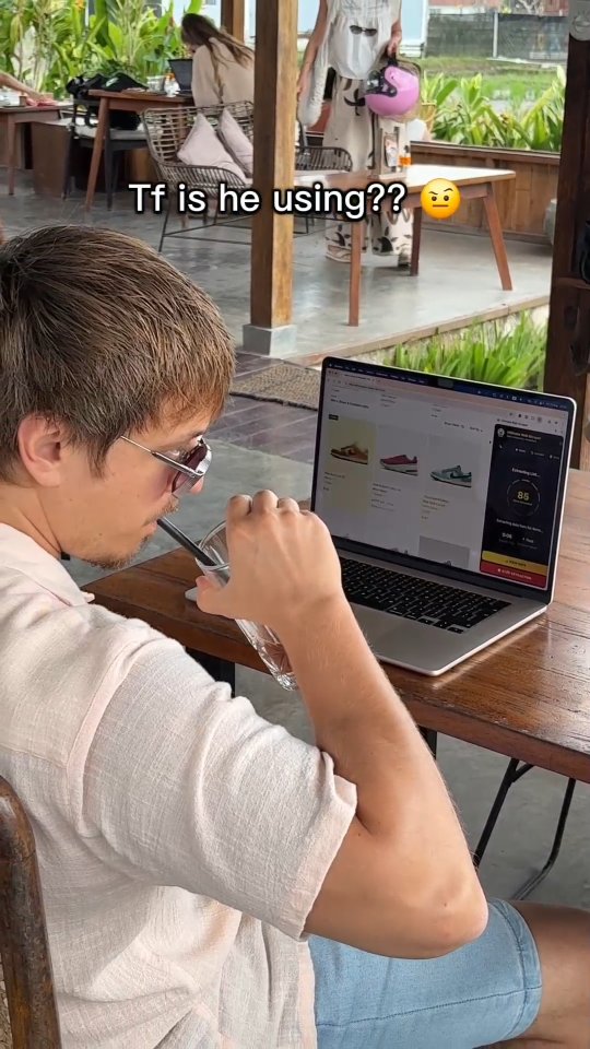 A man sits at a table, looking at a laptop. The laptop screen displays a website with images of shoes. He holds a glass with a straw in his hand while looking at the laptop. The background shows an outdoor cafe setting with other people.