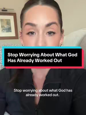 A woman with light skin and dark hair looks directly at the camera. She is wearing a black top. Overlayed on the image is a text box with the words 'Stop Worrying About What God Has Already Worked Out'. The background is a room with a framed picture.