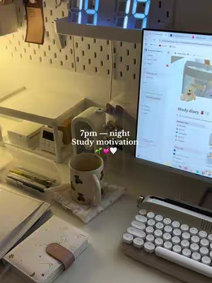 A well-lit desk setup with a computer monitor displaying a Notion page. The desk features a keyboard, mouse, coffee mug, notebooks, and pens. Decorative elements include a digital clock and a string light. The overall aesthetic is clean and minimalist.