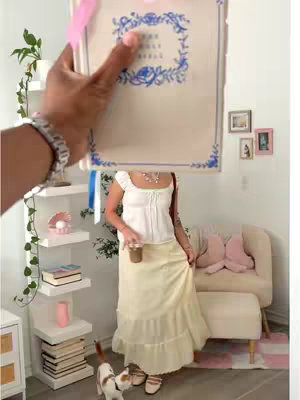 A person holding a bible in front of them, standing in a well-lit room. The person is wearing a white top and long yellow skirt. The scene includes a beige couch, shelves with items on them, and a cat nearby.
