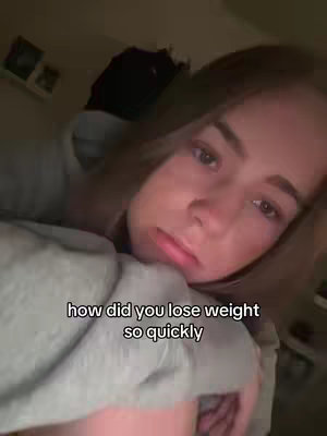 A young woman with brown hair is lying down, appearing to be in bed. A text overlay asks how she lost weight quickly. The image is softly lit, with a blurred out background, suggesting an intimate setting.